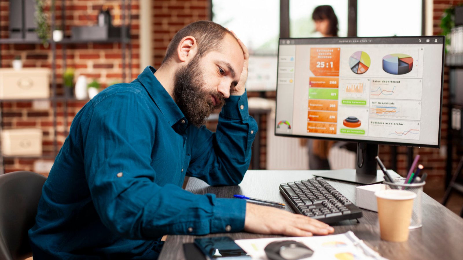 Tired businessman sitting at desk and holding his forehead in startup office. Exhausted male employee having stacks of financial paperwork and computer on table, reflecting demanding work schedule.