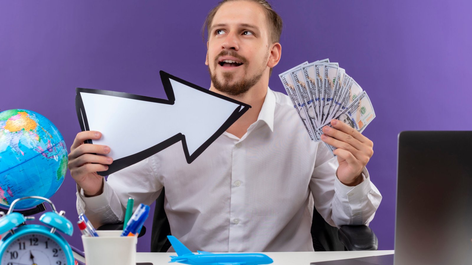 handsome businessman in white shirt holding white arrow showing cash looking aside with smile on face sitting at the table in offise over purple background